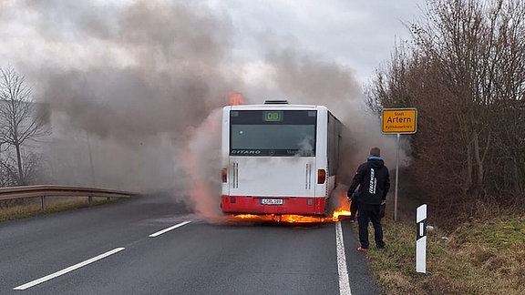 Ein Mann steht hinter einem brennenden Bus neben dem Ortsschilder von Artern