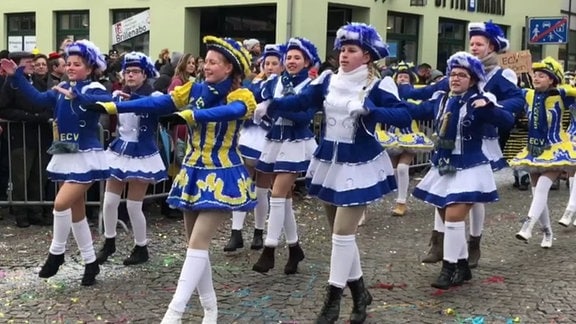 Mädchen in blau-weißen und balu-gelben Karnevalsuniformen tanzen auf Straße vor Zuschauern.