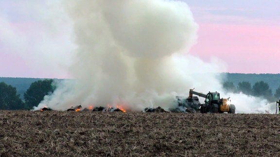 Mehrere brennende Strohballen auf einem Feld werden von einem Bagger zusammengeschoben.