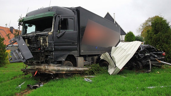 Ein Lkw steht nach einem Unfall auf einem Feld. Di Front ist zerstört. In der Fahrerkabiene fehlen Scheiben. Neben dem Lkw steht ein Auto. Es ist nach einem Zusammenprall zerstört. Auf dem Dach des Autos liegt ein Wellblech.