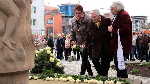 Drei Frauen legen an einem Denkmal weiße Rosen nieder. Hinter ihnen stehen Menschen und schauen zu