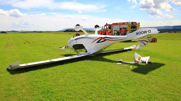 Ein Flugzeug liegt umgedreht und beschädigt auf einer Wiese, im Hintergrund ist ein Feuerwehrauto 
