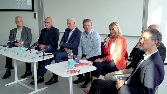 Sechs Männer und eine Frau sitzen bei einer öffentlichen Diskussion nebeneinander.