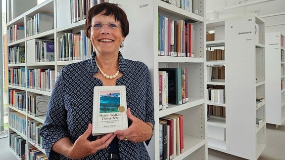 Eine Frau steht vor einem Bücherregal und lacht in die Kamera. Sie hält ein Buch in den Hände. Auf dem Buch steht Martin Walker: "Tête-à-Tête". 