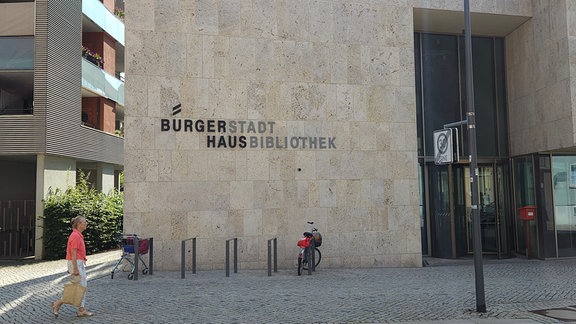 Ein Bau aus Sichtbeton mit den Aufschriften "Bürgerhaus und Stadtbibliothek" ist zu sehen. Davor läuft ein Frau und ein Fahrrad ist an einem Ständer angeschlossen