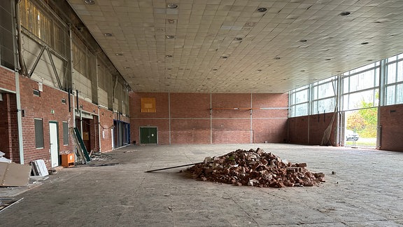 Eine alte entkernte Turnhalle. In der Mitte der großen Halle liegt auf dem Steinboden ein Haufen Schutt.