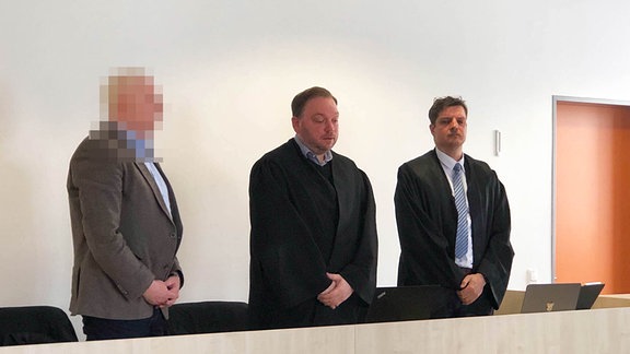 Drei Männer stehen in einem Gerichtssaal.