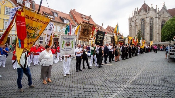 Vertreterinnen und Vertreter unterschiedlicher Kirmesgemeinden stehen aufgereiht auf dem Marktplatz in Mühlhausen. Sie tragen teils Trachten und schwenken bunten Wappen.