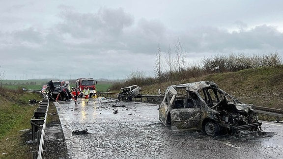 Nach einem Unfall liegen drei Autos auf der B247 bei Bad Langensalza, zwei davon sind ausgebrannt. Im Hintergrund sind Helfer von Feuerwehr und Rettungsdienst im Einsatz.