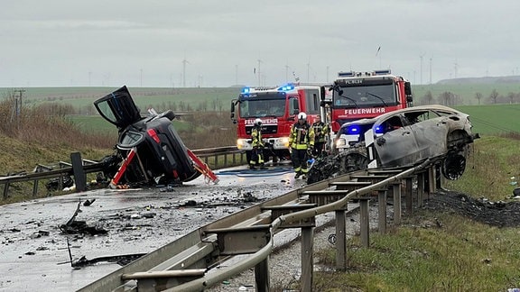 Nach einem Unfall liegen zwei Autos auf den Leitplanken der B247 bei Bad Langensalza, eines davon ist ausgebrannt. Im Hintergrund sind Helfer von Feuerwehr und Rettungsdienst im Einsatz.