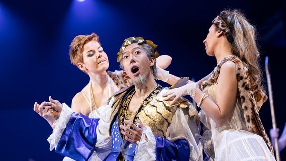 Ein Ensemblemitglied des Theaters Altenburg-Gera steht in fantasievoller, festlicher Kleidung singend auf einer Bühne, daneben zwei weitere Darstellerinnen in weißer Spitze gekleidet.