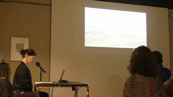 Eine Frau sitzt an einem Tisch mit aufgeklapptem Laptop und Mikrofon und spricht zu einer an die Wand geworfenen Präsentation