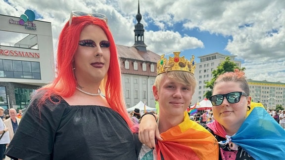 Drei Menschen stehen nebeneinander: Eine Dragqueen, ein junger Mann mit einer Krone auf dem Kopf und eine Frau.