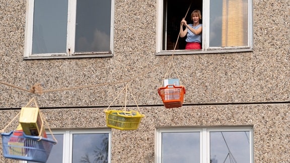 Eine Frau steht am offenen Fenster und hält eine Leine, an der mehrere Wäschekörbe mit Papkartons hängen.