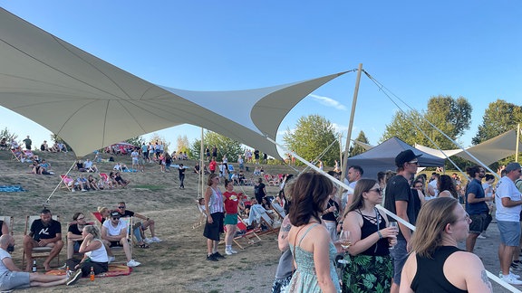 Das Gelände des "Geht Raus"-Open Air in Gera liegt an einem kleinen Wiesenhang. Darauf sitzen und chillen viele Menschen. Manche haben es sich unter einem Sonnensegel bequem gemacht. Zur Bühne hin stehen und tanzen Menschen. 