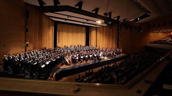 Ein großes Orchester steht auf einer Bühne in einem großen Saal.