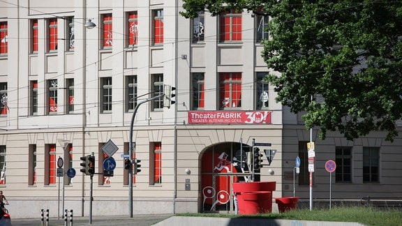 Ein Eckgebäude an einer Straßenkreuzung ist mit einem Banner mit der Aufschrift "Theaterfabrik 30, Theater Altenburg Gera" über dem Eingang behangen. Die Fensterscheiben sind mit rot-weißen Comicmotiven gestaltet.