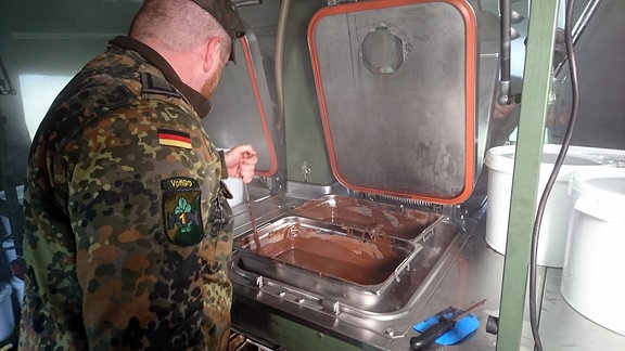 Ein Soldat in Uniform rührt in einer Großküche in einem großen Zelt flüssige Schokolade