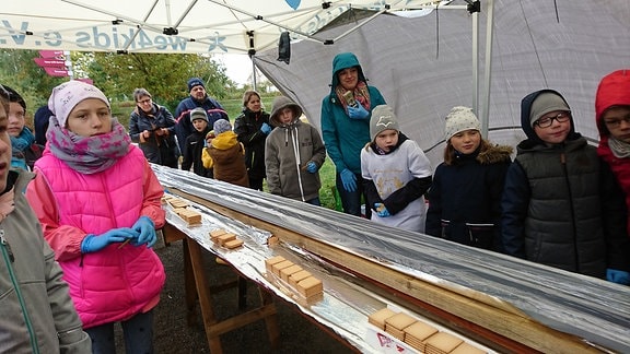 Zahlreiche Kinder stehen neben einem Tisch, auf dem eine mit Alufolie ausgeschlagene Dachrinne und Kekse liegen
