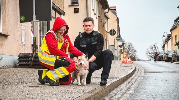 Eine Frau, ein Polizist und ein Hund hocken auf einem Fußweg vor einem Gebäude.