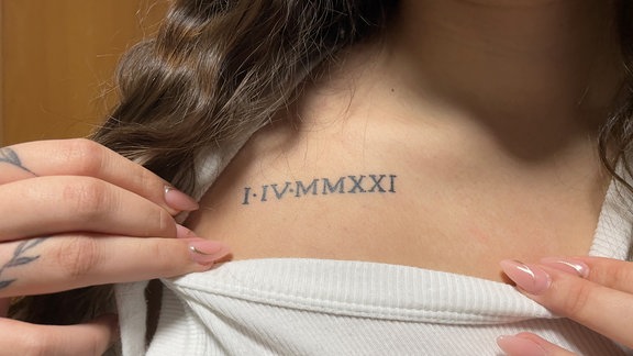 Ein Tattoo mit der Schrift "I.IV.MMXXI" auf dem Schlüsselbein einer Frau