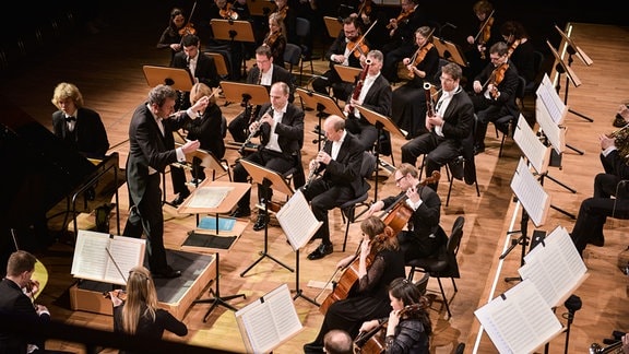 Musikerinnen und Musiker der Jenaer Philharmonie sitzen mit ihren Instrumenten in einem Halbkreis auf einer Bühne.