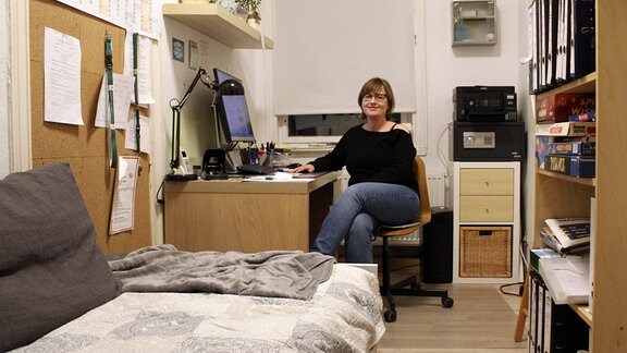 Die Teamleiterin, eine Frau mit Brille und schwarzem Pulli im Porträt, an ihrem Schreibtisch in ihrem Büro mit einem Bett