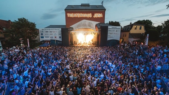 Eine feiernde Menschenmenge vor einer Bühne, dahinter ist das Theaterhaus Jena zu sehen.