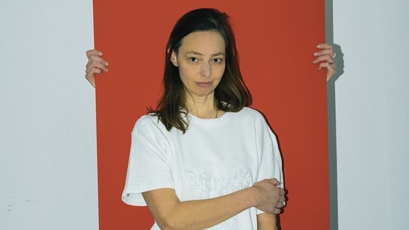 Eine Frau mit langem, offenem Haar trägt ein weißes T-Shirt und blaue Jeans. Sie steht vor einem orangenen Rechteck, das von einer anderen verdeckten Person, von der nur die Hände sichtbar sind, gehalten wird.