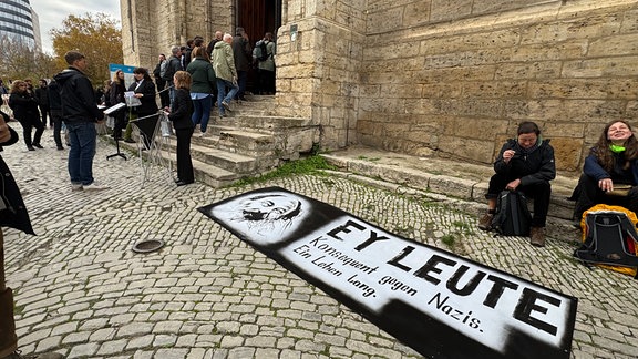 Menschen vor einer Kirch. Auf dem Weg liegt ein großes Schwarzes banner. Drauf ist das Gesicht von Lothar König und die Sätze: "Ey leute, konsequent gegen Nazis. Ein Leben lang".