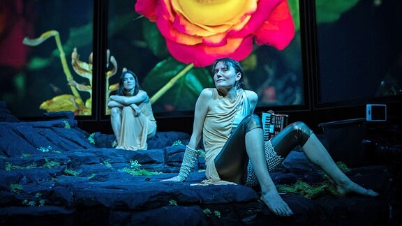 Mystic Vibes, eine große projizierte Blüte mit einem Auge in der Mitte, davor zwei Frauen auf einer Theaterbühne.