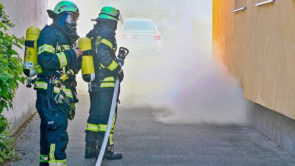 Zwei Feuerwehrleute stehen vor Rauch bei einer Hauswand.