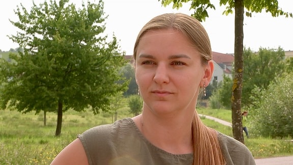 Eine junge Frau (Khrystyna Zhuk) im Park. Sie schaut ernst in die Kamera. Sie trägt ein olivgrünes Sommerkleid und hat die Haare zu einem Pferdeschwanz zusammengebunden.