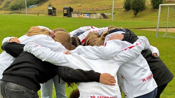 Eine Mädchenfußballmannschaft steht auf einem Fußballplatz im Kreis und steckt die Köpfe zusammen.