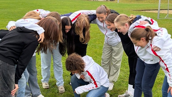 Eine Mädchenfußballmannschaft steht auf einem Fußballplatz im Kreis und macht einen Schlachtruf. Ein Mädchen kniet in der Mitte.