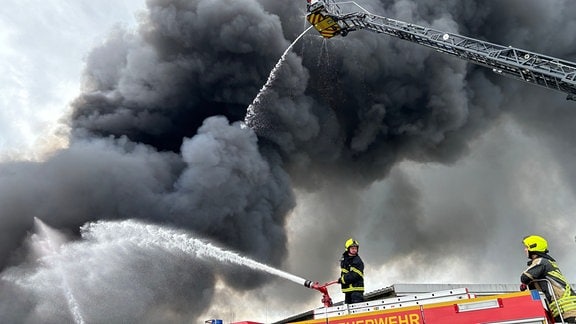 Feuerwehrleute auf einem Feuerwehrauto vor einer großen Rauchwolke. Sie spritzen Wasser aus Schläuchen.
