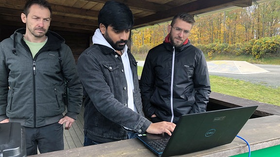 Drei Männer schauen auf einen Laptop. 