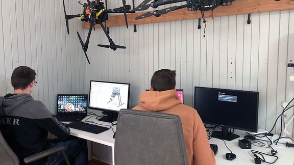 Zwei Männer arbeiten am Computer, über ihnen hängen Drohnen.