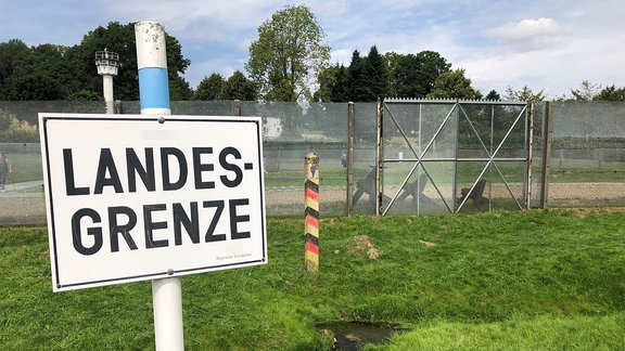 Ein Schild mit der Aufschrift "Landesgrenze" steht vor einem hohen Zaun. 