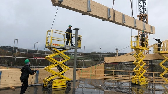 Bauarbeiter auf Hebebühnen helfen einen Holzträger zu instalieren.