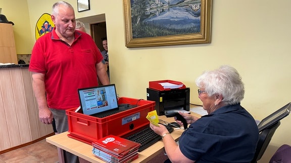 Ein älterer Mann steht vor einem Tisch auf dem Geräte und Blutspende-Broschüren stehen. Eine ältere Frau auf der anderen Seite des Tisches scannt einen Blutspendepass.