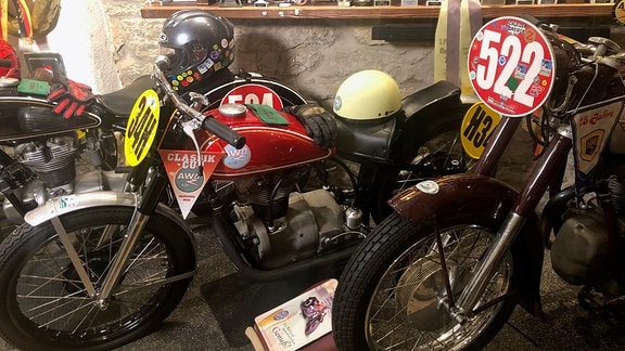 Zwei historische Motorräder, im Hintergrund stehen mehrere Pokale auf einem Regalbrett. 