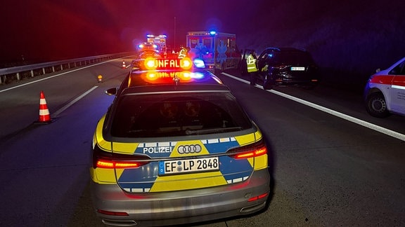 Ein kaputtes Unfallauto auf einer Auto. Rettungskräfte von Feuerwehr und Polizei stehen um das Auto.