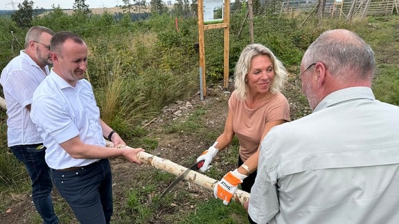 Thüringens Agrarministerin Susanna Karawanskij zersägt einen dicken Holzpfahl, der von Landrat Christian Herrgott und zwei weiteren Männern gehalten wird.