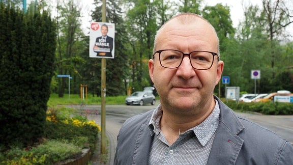 Thomas Benninghaus auf einer Straße in Rudolstadt. Im Hintergrund hängt ein Wahlplakat der Alternative für Saalfeld-Rudolstadt. Benninghaus hat kurze Haar, trägt Brille und ein graues Sakko.