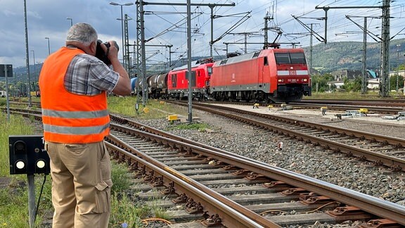 Ein Mann mit einer Warnweste steht neben einem Gleis und fotografiert einen einfahrenden Zug.