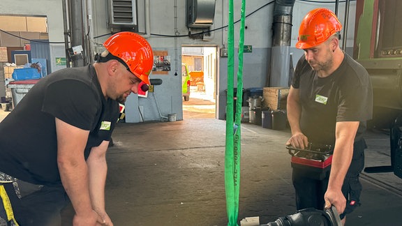 Zwei behelmte Mitarbeiter der Salzburger Eisenbahn-Transportgesellschaft. Zwischen ihnen hängt ein schweres Bauteil an einem Seil.