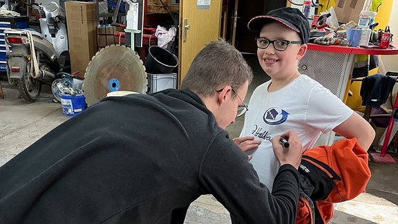 Ein junger Mann unterschreibt auf einem T-Shirt bei einem kleinen Jungen.
