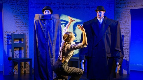 Zwei Schauspielerinnen und ein Schauspieler bespielen eine in blaues Licht getauchte Theaterbühne.
