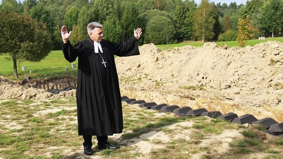 Michael Schwarzkopf, Pastor der Petersburger Petrikirche, steht neben schwarzen Pappsärgen in einem von einem Bagger ausgehobenen Graben auf dem Soldatenfriedhof Sologubowka bei St. Petersburg.